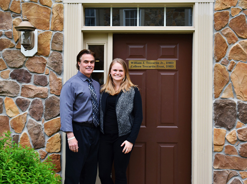 A man and a woman stand together in front of a door with a sign above it, both smiling at the camera.