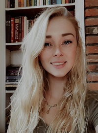 A young woman with blonde hair wearing a necklace is smiling at the camera while posing indoors.