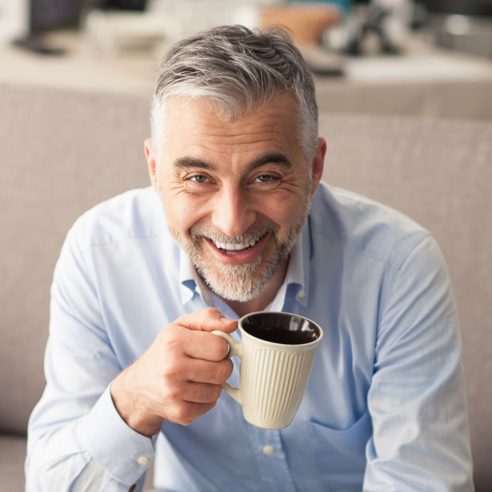 A man with gray hair and a beard is smiling while holding a coffee mug.